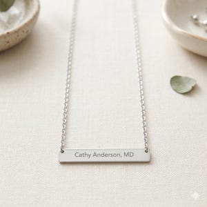 Puede incluir: Un collar plateado con una cadena fina. El colgante rectangular está grabado con el texto "Cathy Anderson, MD". El collar se muestra sobre una superficie blanca, con pequeños cuencos decorativos en el fondo.