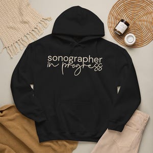 May include: A black hooded sweatshirt featuring the text "sonographer in progress" printed in a light beige color across the chest. The hoodie includes a front kangaroo pocket, long sleeves, and a drawstring hood with matching black drawstrings.
