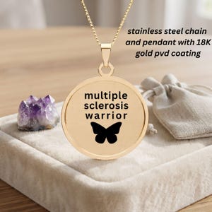 May include: A gold-colored circular pendant on a chain, featuring the text "multiple sclerosis warrior" and a black butterfly silhouette. The pendant is made of stainless steel with an 18K gold PVD coating, presented with a small fabric pouch.