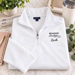 May include: A white quarter-zip pullover featuring a Sport-Tek brand label at the inner collar. The chest displays black text that reads "BEHAVIOR Analyst" with a small heart icon and the name "Linda" printed in a cursive script font below.