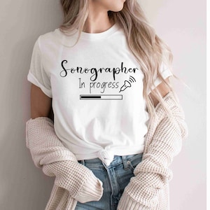 Puede incluir: Camiseta blanca con el texto "Sonógrafo En Progreso" y un gráfico de barra de progreso con un pequeño icono de una sonda de ultrasonido médica.