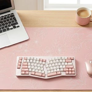 May include: A large rectangular pink desk mat featuring a subtle white line-art floral pattern. The mat is placed on a wooden surface and holds a silver laptop, an ergonomic split keyboard with pink and white keys, a rose gold mouse, and a mug.