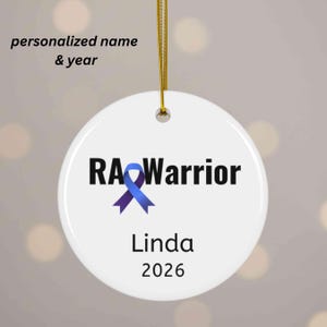 May include: A white, round ornament with the text "RA Warrior" in black, featuring a blue ribbon graphic. Below, the name "Linda" and the year "2026" are printed. A gold string is attached for hanging. The words "personalized name & year" are above the ornament.