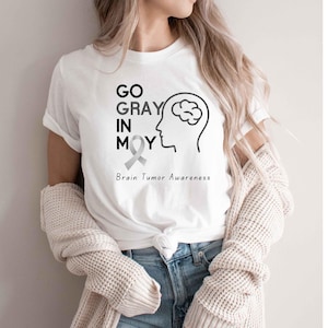 May include: White t-shirt with the text "GO GRAY IN MAY" and a brain illustration. The shirt also includes a gray ribbon and the words "Brain Tumor Awareness" in a simple, clean font. The design is centered on the front of the shirt.
