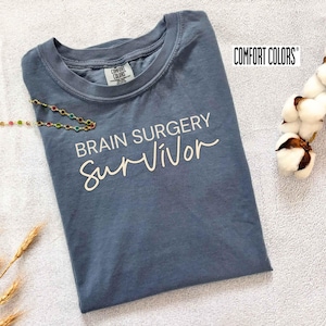 May include: A slate blue Comfort Colors t-shirt with the words "BRAIN SURGERY Survivor" printed in white script. The shirt is folded and lies on a white surface, with a colorful beaded necklace and cotton sprig nearby.
