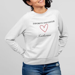 Puede incluir: Una sudadera blanca con el texto "FAVORITE DAUGHTER", un coraz&oacute;n rojo y el nombre "Katherine". La sudadera es de cuello redondo. La persona lleva vaqueros azules.