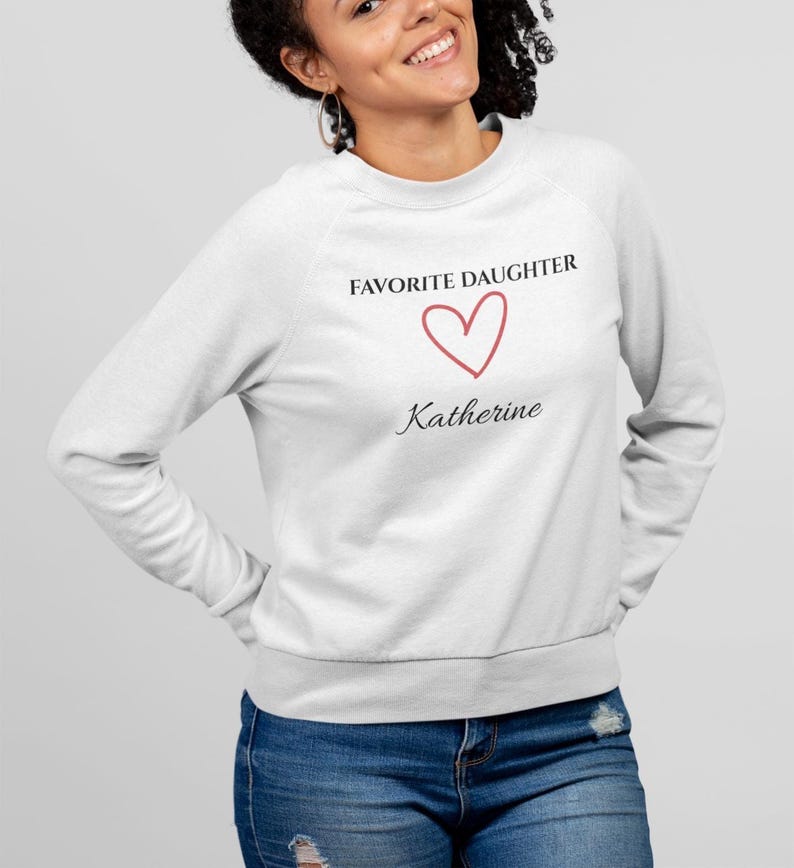 Puede incluir: Una sudadera blanca con el texto "FAVORITE DAUGHTER", un coraz&oacute;n rojo y el nombre "Katherine". La sudadera es de cuello redondo. La persona lleva vaqueros azules.