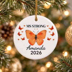 May include: A white circular ceramic ornament featuring a large orange butterfly in the center, surrounded by smaller orange butterflies. The text MS STRONG is printed above the butterfly, with the name Amanda and the year 2026 below it.