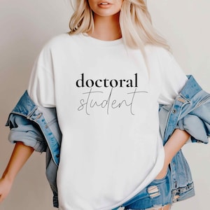 May include: White t-shirt with the words "doctoral student" in black script. The shirt is worn with a denim jacket and blue jeans. The text is in a stylish, modern font.