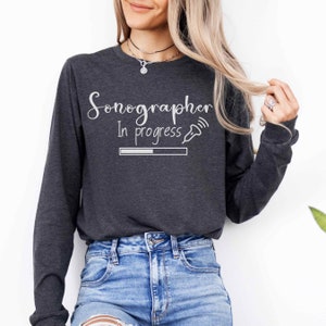 Może przedstawiać: Szary, długos rękawowy t-shirt z napisem "Sonograf w trakcie" i grafiką przedstawiającą medyczną sondę ultradźwiękową z paskiem postępu.