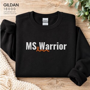 May include: A black crewneck sweatshirt featuring the white text "MS Warrior" printed on the chest. An orange awareness ribbon is integrated into the design, positioned between the letters "MS" and "Warrior". The garment has long sleeves and a ribbed collar.