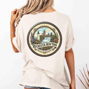 May include: Cream-colored t-shirt featuring a circular graphic of Rochester, New York. The design includes a lighthouse, waterfall, cityscape, and the text "Rochester, New York - Flower City - The Heart of New York".