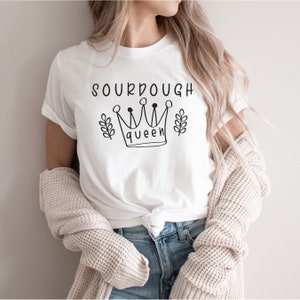 May include: A white short-sleeved t-shirt featuring a black graphic print on the chest. The design includes the text SOURDOUGH in a simple font, a line-drawn crown with the word queen inside, and two small leafy sprigs on either side.