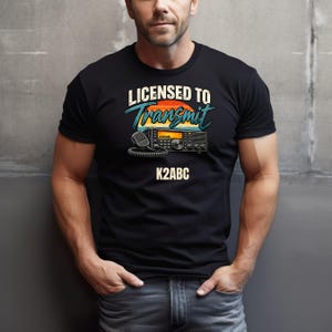 May include: A black short-sleeved t-shirt featuring a graphic print of a radio transceiver with a microphone. Above the radio, the text reads "LICENSED TO Transmit" in a retro font. Below the radio, the alphanumeric text "K2ABC" is printed.