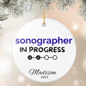 May include: White ceramic ornament with the text "sonographer IN PROGRESS" in blue and black. The ornament has a progress bar with check marks and circles, the name "Madison" and the year "2025".