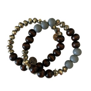 Könnte beinhalten: Zwei Perlenarmbänder, eines mit braunen Holzperlen und das andere mit grauen und goldenen Perlen. Beide Armbänder haben einen kleinen Metallanhänger mit der Aufschrift "Simply Blessed".