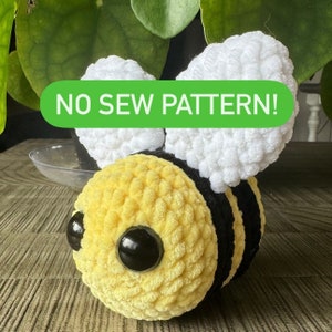 May include: A crocheted bee toy with a yellow and black striped body, white fuzzy wings, and large black eyes. The toy is sitting on a wooden surface. The text "NO SEW PATTERN!" is visible in the image.