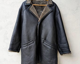 May include: A black leather coat with a shearling lining and collar hangs on a wooden hanger. The coat has two front pockets and a button closure. The shearling lining is visible at the collar, cuffs, and hem.