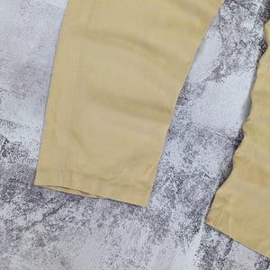 May include: A pair of light tan trousers featuring a straight-leg cut with visible vertical seams along the sides. The fabric has a smooth, matte texture and a consistent solid color throughout the garment, showcasing the lower leg portion of the pants.