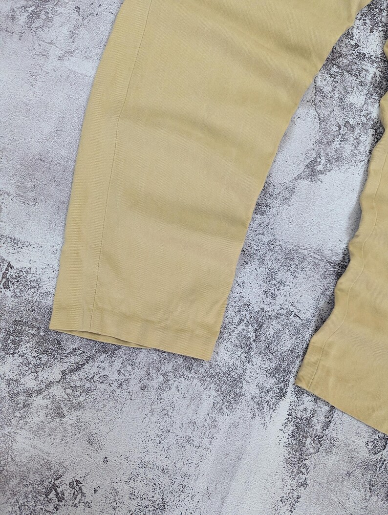 May include: A pair of light tan trousers featuring a straight-leg cut with visible vertical seams along the sides. The fabric has a smooth, matte texture and a consistent solid color throughout the garment, showcasing the lower leg portion of the pants.