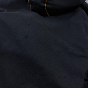 Pode incluir: Uma camisola com capuz preta de tecido texturizado com um fino debrum laranja contrastante ao longo das costuras do capuz. A pe&ccedil;a apresenta pequenos fios puxados vis&iacute;veis na superf&iacute;cie frontal, indicando desgaste no material escuro.