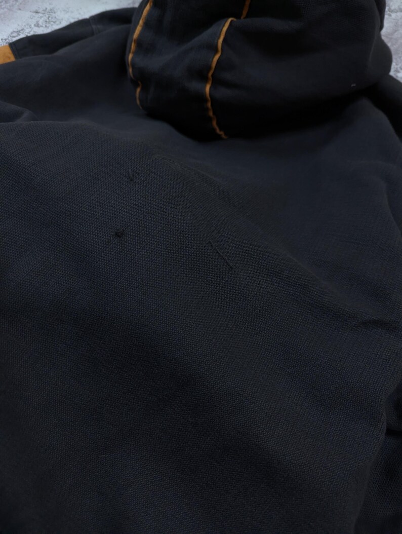 Pode incluir: Uma camisola com capuz preta de tecido texturizado com um fino debrum laranja contrastante ao longo das costuras do capuz. A pe&ccedil;a apresenta pequenos fios puxados vis&iacute;veis na superf&iacute;cie frontal, indicando desgaste no material escuro.