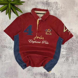 以下が含まれることがあります： 赤色のポロシャツ。青色の数字4と「joules Elephant Polo」の刺繍入り。襟は黄色で、サイドパネルは青色。右側には紋章が刺繍されています。