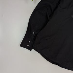 Puede incluir: Camisa negra de manga larga con cuello y pu&ntilde;os abotonados. La camisa presenta un peque&ntilde;o parche con la bandera austriaca en la manga superior. La tela parece ser un material liso y tejido.