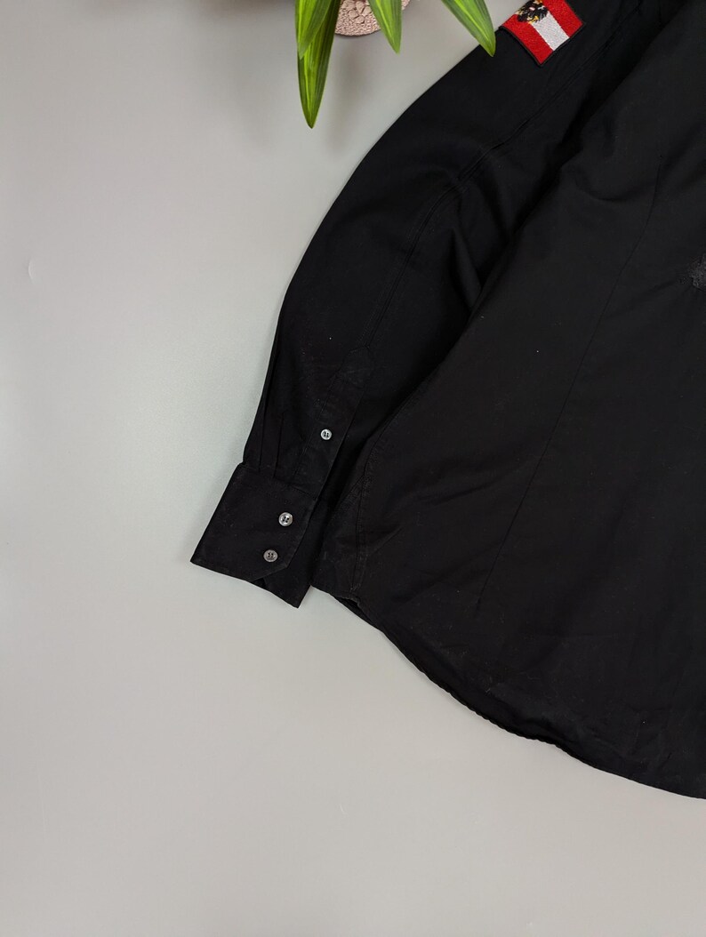 Puede incluir: Camisa negra de manga larga con cuello y pu&ntilde;os abotonados. La camisa presenta un peque&ntilde;o parche con la bandera austriaca en la manga superior. La tela parece ser un material liso y tejido.