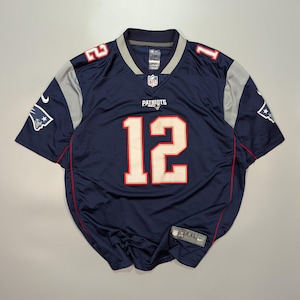Puede incluir: Una camiseta de fútbol azul marino con detalles en blanco y plateado. La camiseta tiene el número "12" en la parte delantera y la palabra "PATRIOTS" encima. El logotipo de la NFL es visible en el pecho. La camiseta es talla XXL.