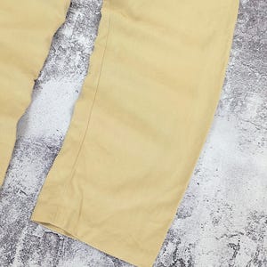 May include: A pair of light tan trousers featuring a straight-leg cut with visible vertical seams running down the front of each leg. The fabric has a smooth, matte finish and a consistent solid color throughout the garment, showing a clean hem.