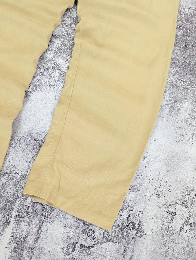 May include: A pair of light tan trousers featuring a straight-leg cut with visible vertical seams running down the front of each leg. The fabric has a smooth, matte finish and a consistent solid color throughout the garment, showing a clean hem.