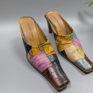 May include: A pair of patchwork leather mules with a square toe. The open-back shoes feature a variety of colors, including brown, yellow, pink, and blue. The heels are a dark brown color, and the brand name "Giannmarco Lorenzi" is visible inside the shoe.