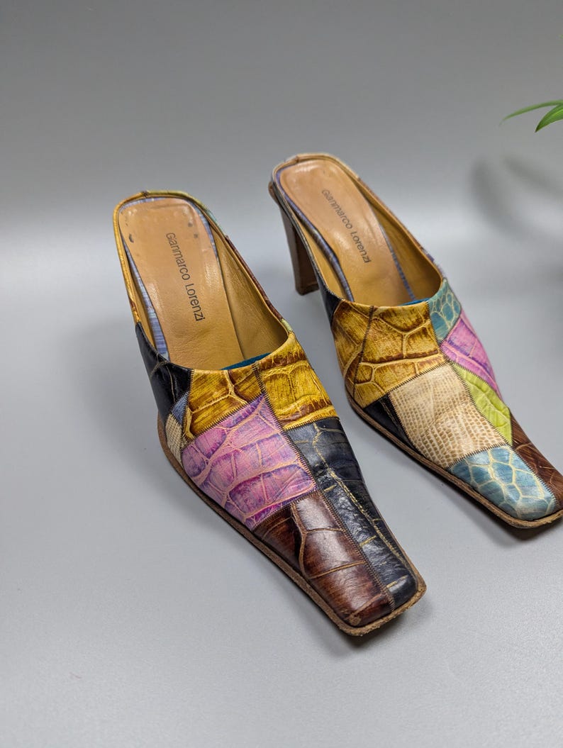 May include: A pair of patchwork leather mules with a square toe. The open-back shoes feature a variety of colors, including brown, yellow, pink, and blue. The heels are a dark brown color, and the brand name "Giannmarco Lorenzi" is visible inside the shoe.