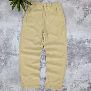 May include: A pair of solid beige trousers featuring an elasticized waistband, a relaxed fit through the legs, and two side pockets. The pants are constructed with a simple, clean design and a smooth, uniform fabric texture throughout the garment.