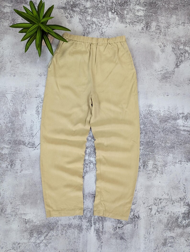 May include: A pair of solid beige trousers featuring an elasticized waistband, a relaxed fit through the legs, and two side pockets. The pants are constructed with a simple, clean design and a smooth, uniform fabric texture throughout the garment.