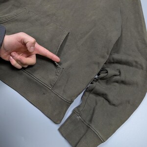 Puede incluir: Sudadera con capucha Carhartt verde oliva con bolsillo delantero y el nombre de la marca impreso en negro. La sudadera tiene una capucha con cord&oacute;n y pu&ntilde;os y dobladillo acanalados. La tela parece desgastada y ligeramente descolorida.