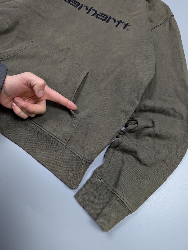 Puede incluir: Sudadera con capucha Carhartt verde oliva con bolsillo delantero y el nombre de la marca impreso en negro. La sudadera tiene una capucha con cord&oacute;n y pu&ntilde;os y dobladillo acanalados. La tela parece desgastada y ligeramente descolorida.
