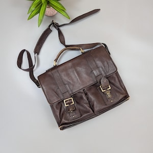 Könnte beinhalten: Eine braune Ledertasche mit einem oberen Griff und einem verstellbaren Schultergurt. Die Tasche hat zwei Vordertaschen, die mit Messingschnallen gesichert sind. Die Tasche ist etwa 30 cm breit und 25 cm hoch.