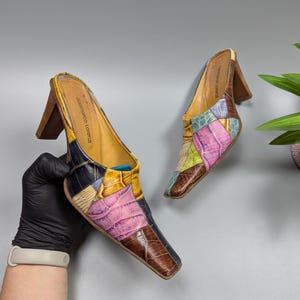 Op de afbeelding: Een paar kleurrijke patchwork leren muiltjes met hak. De schoenen hebben een vierkante neus, houten hakken en een verscheidenheid aan kleuren, waaronder bruin, roze, blauw en geel. De merknaam "Gianmarco Lorenzi" is zichtbaar op de binnenzool.