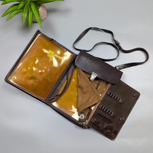 May include: Open brown leather artist's portfolio with a shoulder strap. The portfolio has multiple compartments, including a clear plastic sleeve and a section for holding pencils and tools. The interior is lined with a tan fabric.