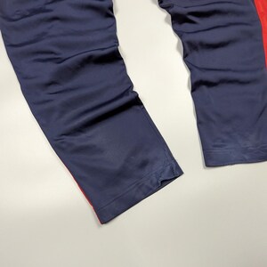 May include: Navy blue athletic trousers with a red stripe down the side. The trousers are made of a smooth, possibly satin-like material. The image shows the lower portion of the trousers, with the hem visible.