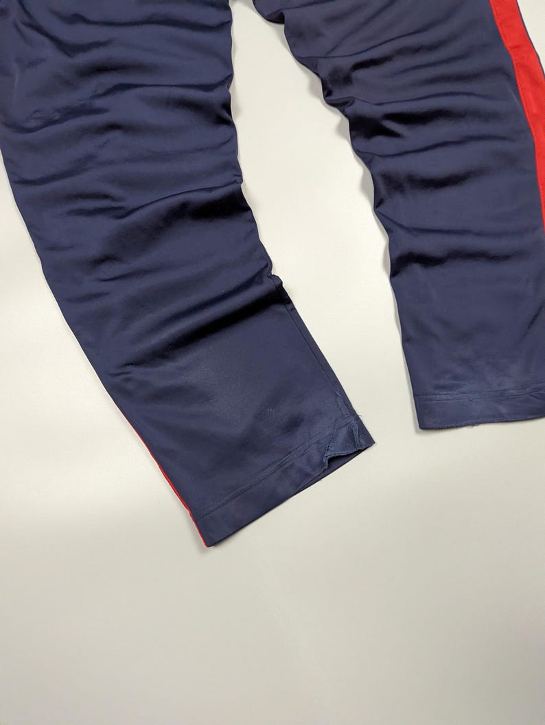 May include: Navy blue athletic trousers with a red stripe down the side. The trousers are made of a smooth, possibly satin-like material. The image shows the lower portion of the trousers, with the hem visible.