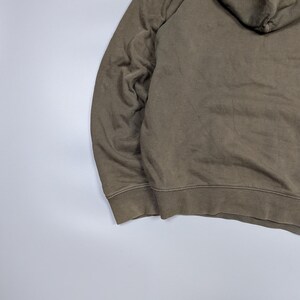 Puede incluir: Una sudadera con capucha verde oliva descolorida con capucha con cord&oacute;n y pu&ntilde;os acanalados. La sudadera est&aacute; hecha de un material suave y desgastado, lo que sugiere comodidad y uso informal. La imagen muestra detalles de la sudadera.