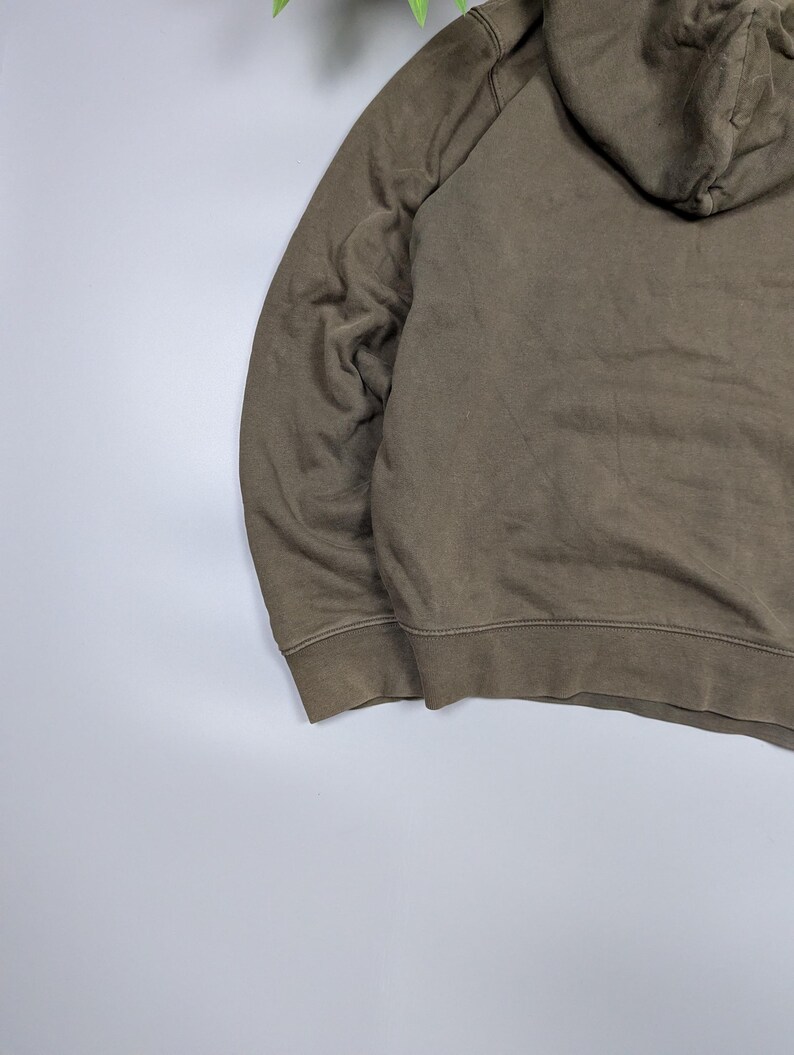 Puede incluir: Una sudadera con capucha verde oliva descolorida con capucha con cord&oacute;n y pu&ntilde;os acanalados. La sudadera est&aacute; hecha de un material suave y desgastado, lo que sugiere comodidad y uso informal. La imagen muestra detalles de la sudadera.