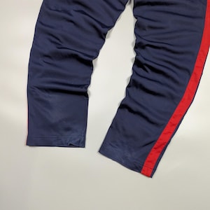 May include: Navy blue tracksuit bottoms with a bold red stripe running down the side. The trousers appear to be made from a smooth, possibly satin-like fabric. The legs are straight and slightly creased.