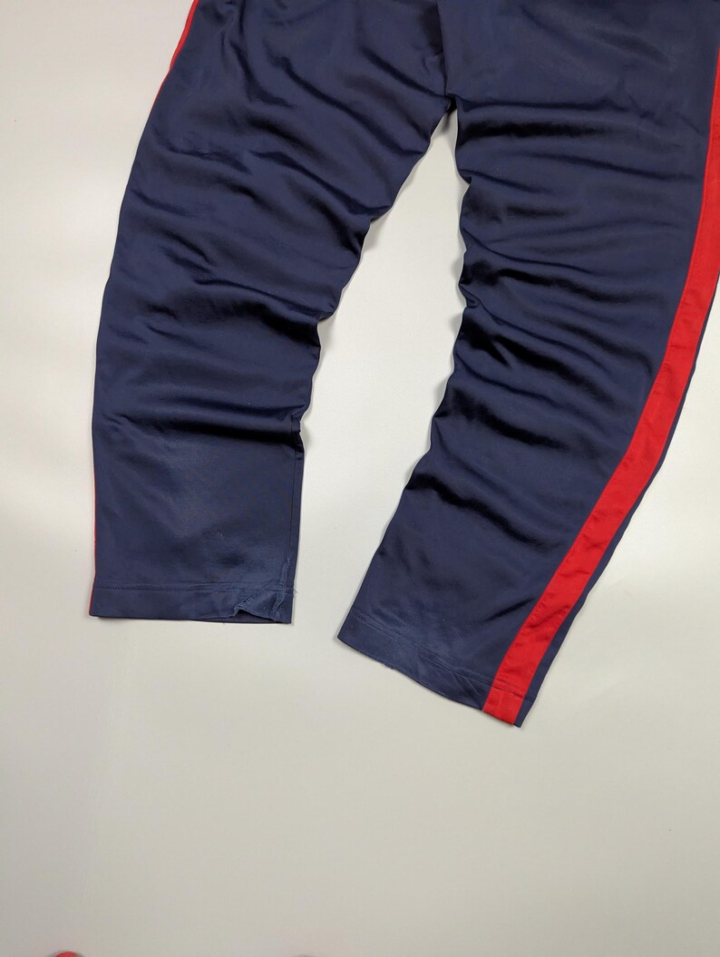 May include: Navy blue tracksuit bottoms with a bold red stripe running down the side. The trousers appear to be made from a smooth, possibly satin-like fabric. The legs are straight and slightly creased.