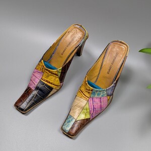 May include: A pair of patchwork leather mules with square toes. The shoes feature a variety of colors, including brown, yellow, pink, and blue. The heels are a dark brown color, and the interior is tan. The brand name "Gammarco Lorenzi" is visible inside the shoe.