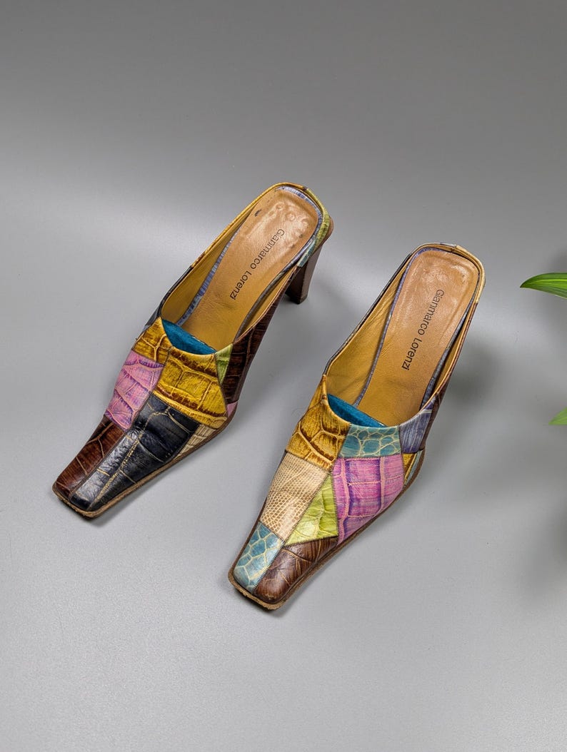 May include: A pair of patchwork leather mules with square toes. The shoes feature a variety of colors, including brown, yellow, pink, and blue. The heels are a dark brown color, and the interior is tan. The brand name "Gammarco Lorenzi" is visible inside the shoe.