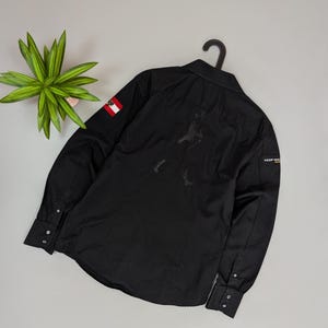 Puede incluir: Camisa negra de manga larga con botones, un dise&ntilde;o gr&aacute;fico sutil en la espalda y un peque&ntilde;o parche de bandera en la manga. Cuello cl&aacute;sico y pu&ntilde;os abotonados. El texto "KEEP WALKING" es visible en la manga.